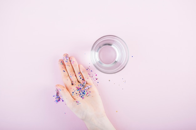 Блестки и блестящая капсула на человеческой руке возле стакана воды на розовом фоне