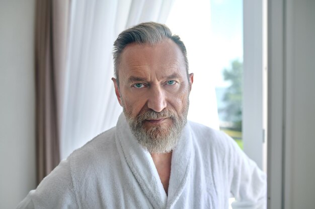 Serene mature gentleman in a white terry bathrobe standing indoors