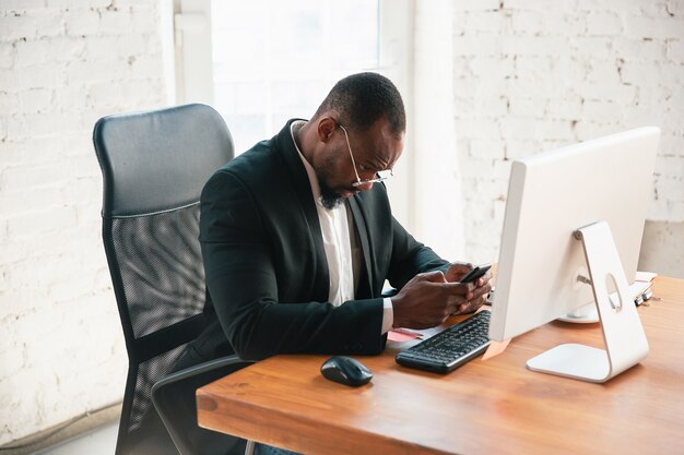 Serfing online. African-american entrepreneur, businessman working concentrated in office. Looks serios and busy, wearing classic suit, jacket. Concept of work, finance, business, success, leadership.