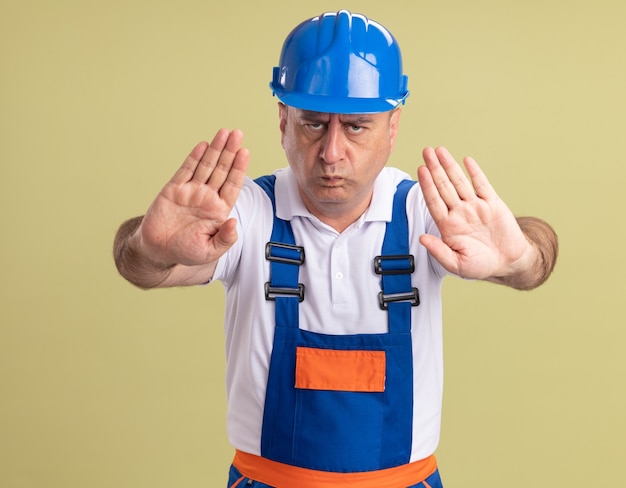 Серьезный взрослый человек-строитель в униформе жестов стоп знак рукой с двумя руками, изолированными на оливково-зеленой стене