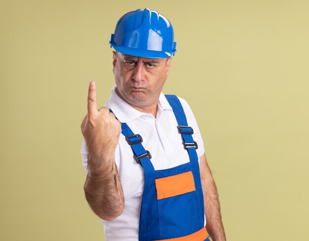 Serious adult builder man in uniform points up isolated on olive green wall