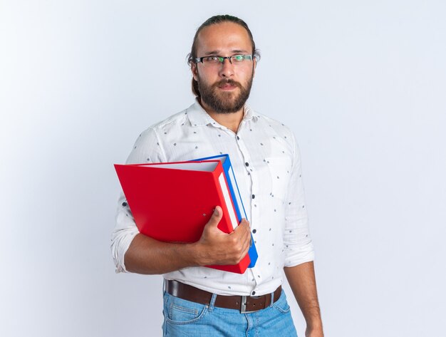 Serious adult handsome man wearing glasses holding folders looking at camera isolated on white wall