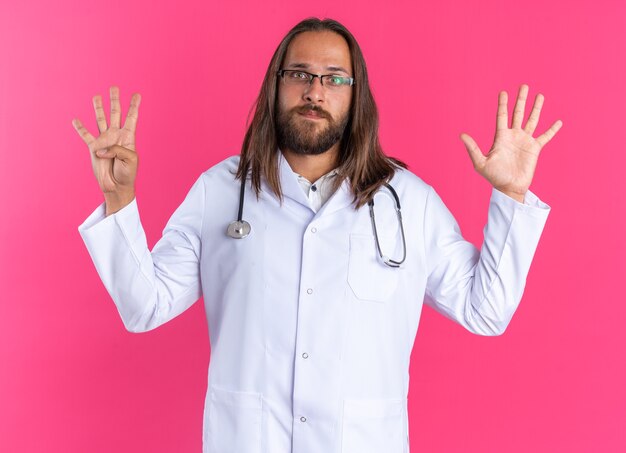 Serious adult male doctor wearing medical robe and stethoscope with glasses looking at camera showing nine with hands isolated on pink wall