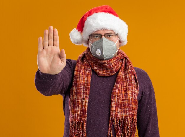 Serious adult man wearing protective mask glasses and santa hat with scarf around neck looking at camera doing stop gesture isolated on orange background