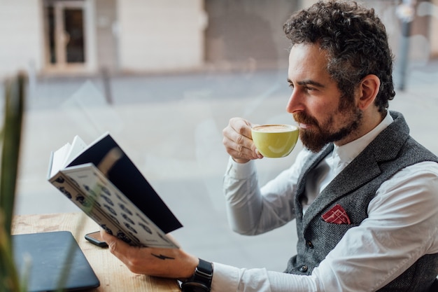 Grave uomo adulto di mezza età sorseggia e gode di una bevanda al caffè durante la lettura di un libro interessante e accattivante nel bar o nella libreria