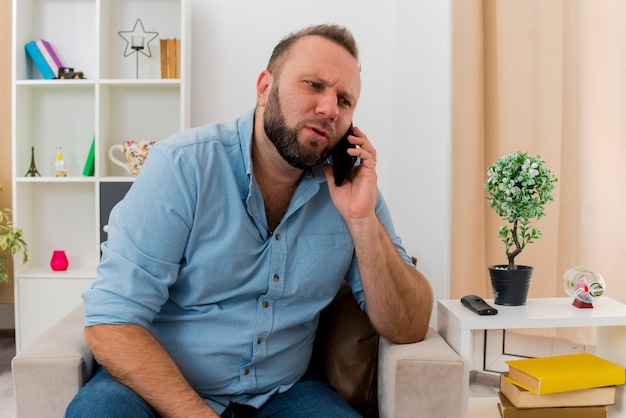 Serious adult slavic man sits on armchair talking on phone inside the living room