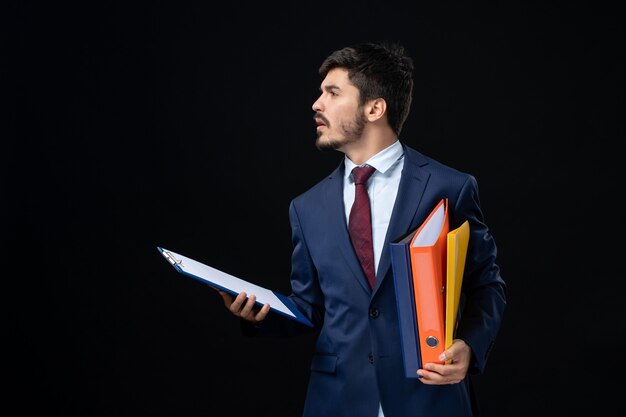 Serious adult in suit holding several documents and looking somewhere on isolated dark wall