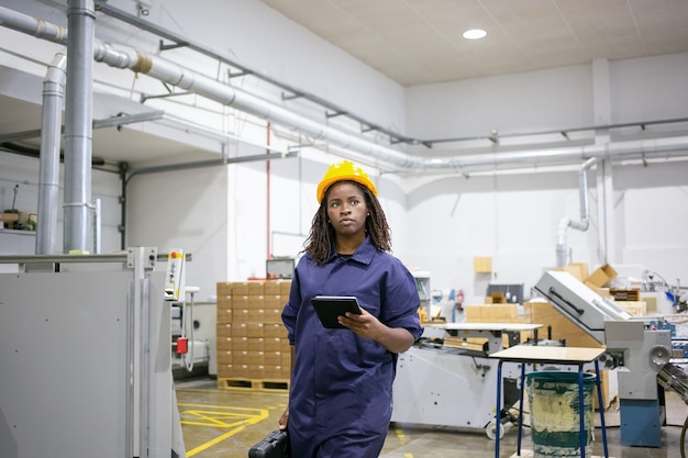 Serious African American female worker in protective uniform walking to workplace on plant floor, holding tablet and case with tools