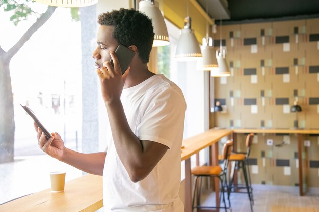 Serious African American guy talking on cell and looking at tablet screen