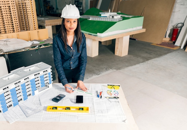 Serious African-American lady in safety helmet standing near model of building on table