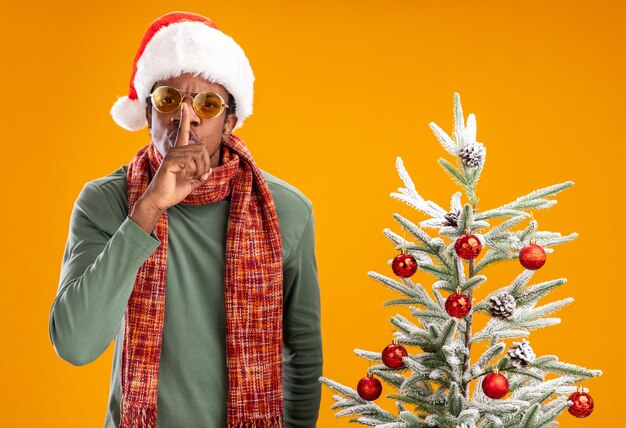 Serious african american man in santa hat and scarf around neck making silence gesture with finger on lips standing next to a christmas tree over orange background