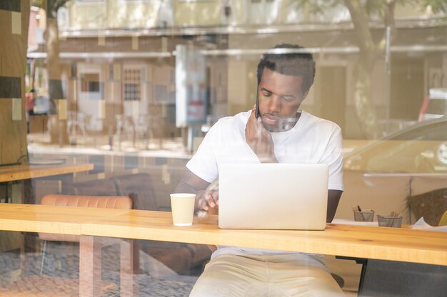 Serious African American man typing on laptop and speaking on cellphone in co-working space