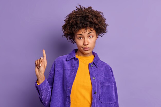 Serious African American woman has curly hair assures you to go upstairs indicates with index finger above wears fashionable clothes.