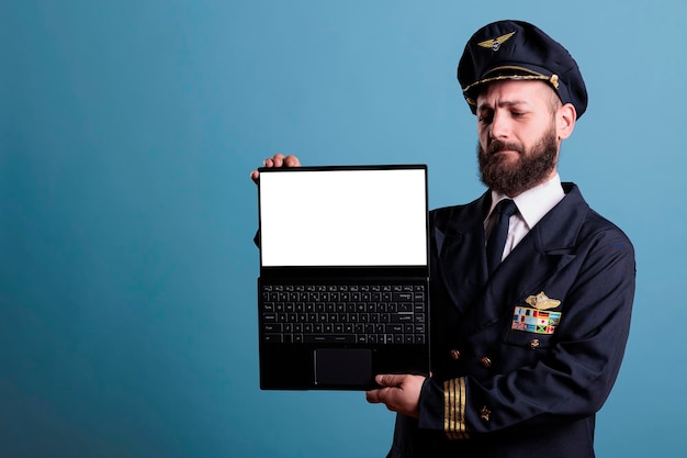 빈 흰색 화면이 있는 노트북, 항공 아카데미 소프트웨어 광고, 공항 웹사이트 모형이 있는 PC를 보여주는 진지한 비행기 조종사. 빈 디스플레이가 있는 휴대용 컴퓨터를 들고 있는 비행기 기장