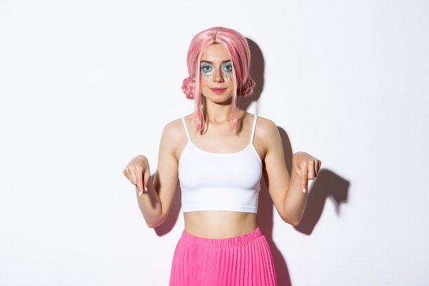 Бесплатное фото Серьезная и уверенная в себе кавказская женщина в розовом аниме-парике и праздничном макияже, указывая пальцами вниз, показывает рекламу о праздновании праздника
