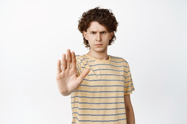 Il ragazzo serio e arrabbiato allunga la mano mostrando il gesto di arresto tabù proibisce lo smth in piedi su sfondo bianco
