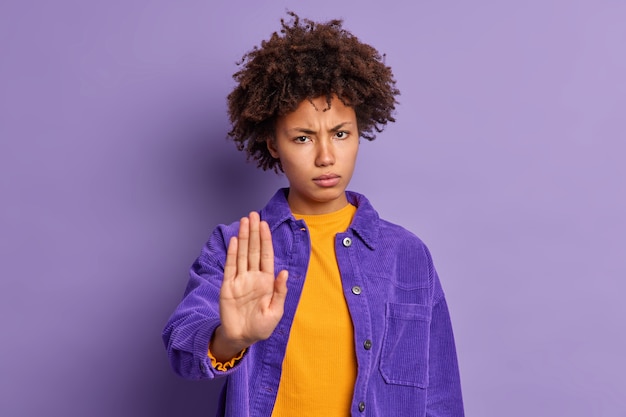 Serious annoyed dark skinned afro american woman keeps palm in stop gesture asks not to bother her looks angrily wears purple jacket expresses restriction or denial. do not come closer please