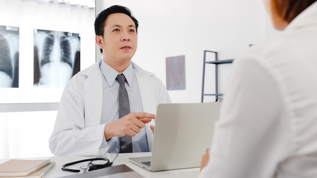 컴퓨터 노트북을 사용하는 흰색 의료 제복을 입은 심각한 아시아 남성 의사가 좋은 소식을 전달하고 있습니다.