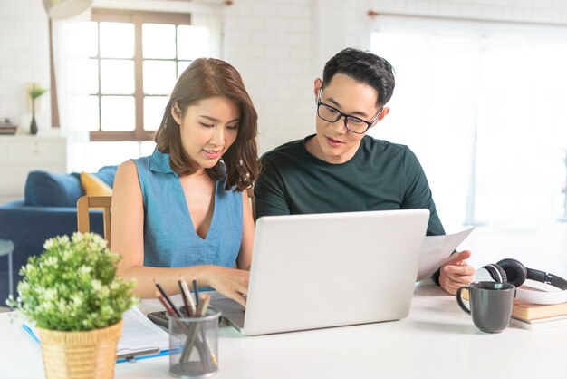 Serious Asian husband checking analyzing statement utilities bills sitting together at home.