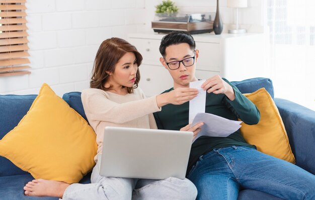 Serious Asian husband checking analyzing statement utilities bills sitting together at home.