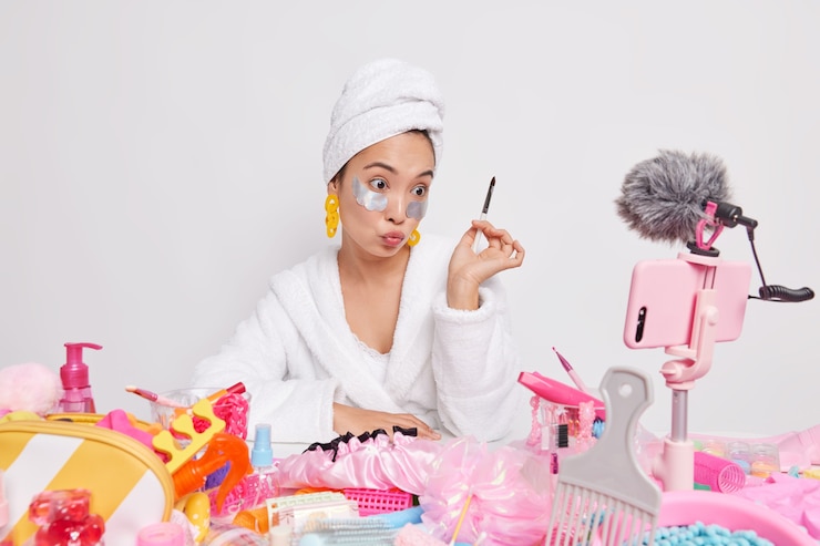 Free Photo serious asian lady beauty bloger records video for her beauty vlog holds cosmetic brush applies eyepatches tells about skin care procedures does makeup at home in front of smartphone camera