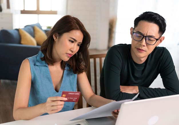 Serious Asian wife checking analyzing statement utilities bills sitting together at home