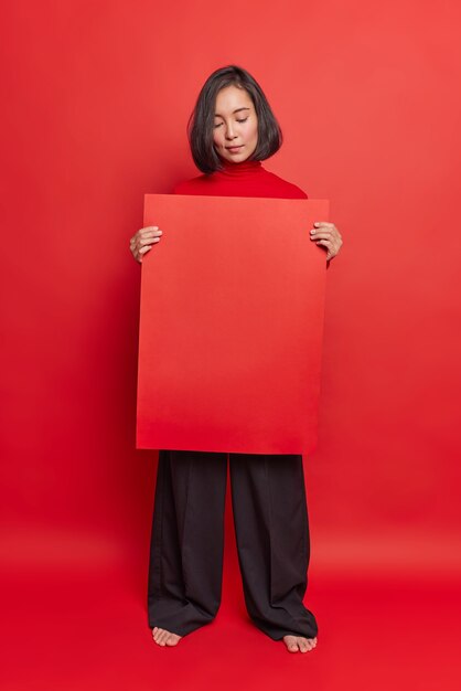 Серьезная азиатская женщина держит красный рекламный щит, рекомендует разместить здесь вашу информацию, демонстрирует баннер в стильном наряде на фоне яркой стены студии.