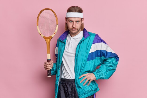 Serious athlete man holds tennis racket dressed in sport outfit looks confidently, poses against pink wall. Unshaven self assured guy going to play badminton. Active life concept