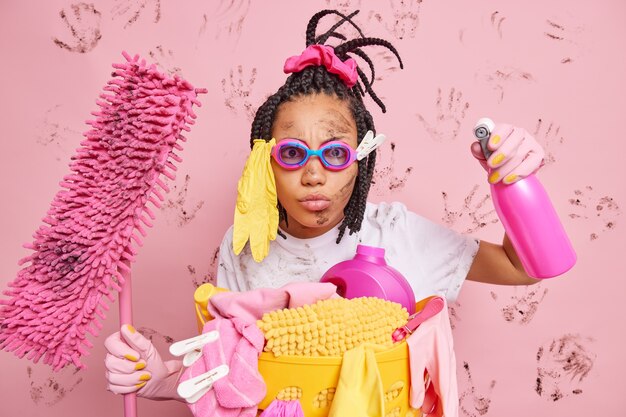 Серьезная внимательная домработница с грязным лицом занята уборкой дома, дезинфицирует комнату со спреем для моющего средства, держит швабру для мытья пола, позирует возле корзины для белья, изолированной над розовой стеной