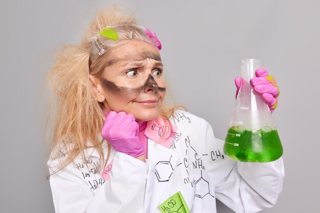 真面目で気配りのある女性薬剤師は、実験が緑色の液体でフラスコを保持し、白衣のゴム手袋を着用し、灰色で分離された表情を驚かせたことを示しています