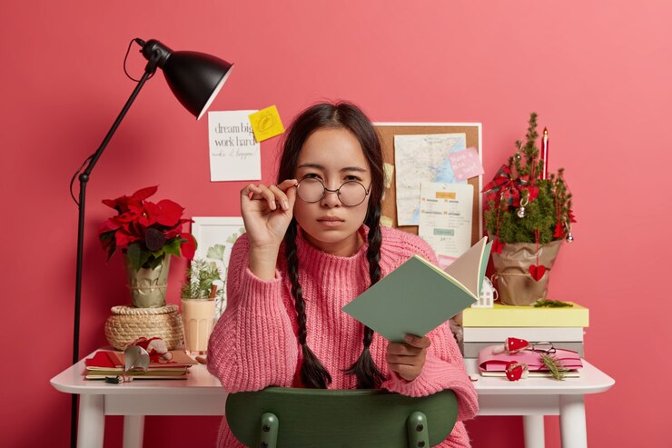 Free Photo serious attractive brunette girl looks suspiciously through round glasses, keeps hand on frame, holds opened notepad or diary