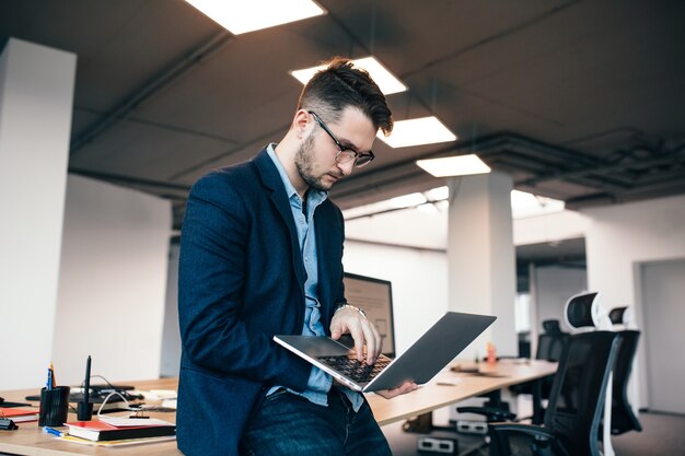 안경에 심각한 매력적인 남자는 사무실에서 직장 근처에 서 있습니다. 그는 파란색 셔츠에 어두운 재킷을 입습니다. 그는 노트북에 입력하고 있습니다.