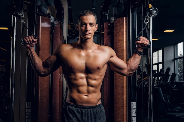 Serious attractive man is doing exercises with training apparatus at dark gym club.