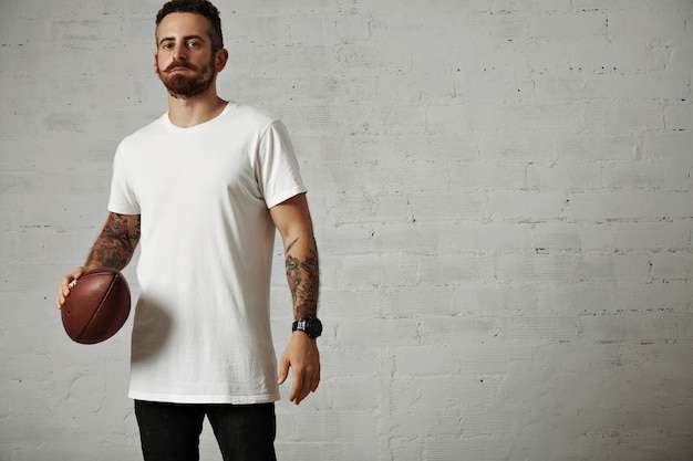 Free photo serious attractive young man in a blank white cotton t-shirt and black jeans holding a vintage rugby ball isolated on white