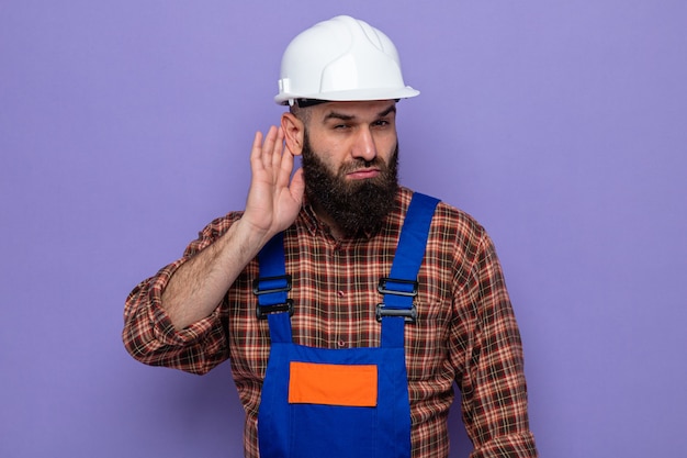 Серьезный бородатый строитель в строительной форме и защитном шлеме пытается слушать, держа руку возле уха