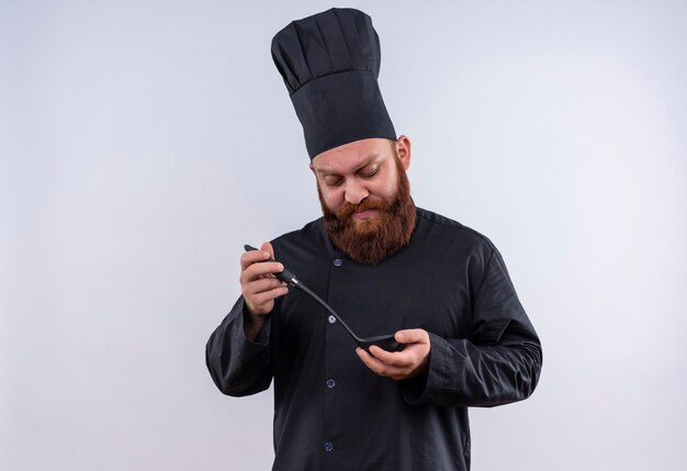 A serious bearded chef man in black uniform holding black ladle and looking at it on a white wall