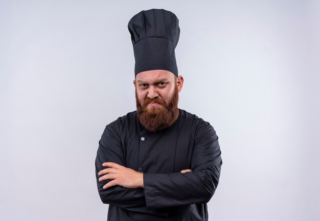 A serious bearded chef man in black uniform holding hands folded while looking on a white wall