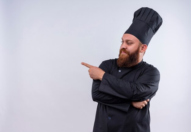 A serious bearded chef man in black uniform pointing left side with index finger on a white wall