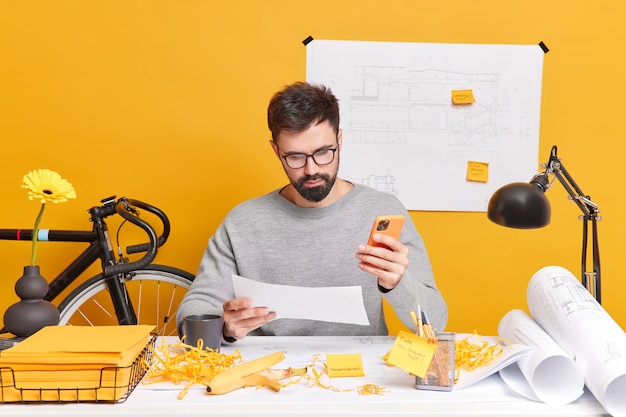 Serious bearded European man concentrated at paper uses mobile phone, makes drawings for construction company surrounded with blueprint rolls stickers and papers. Job concept.