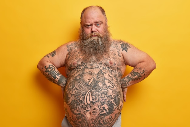 Серьезный бородатый толстяк с густой бородой, татуированным телом и большим животом, смотрит из-под бровей, держит руки на талии, изолирован на желтой стене. Ожирение, липосакция, концепция похудания