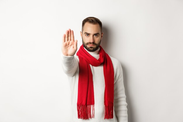Серьезный бородатый парень говорит нет, показывает знак остановки, знак отказа, запрещает действия, стоит в зимнем свитере и красном шарфе на белом фоне