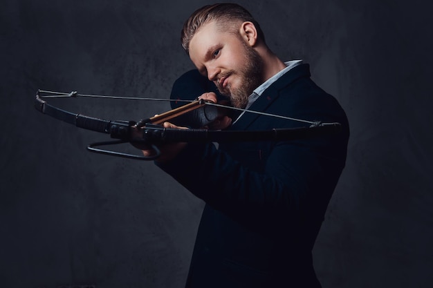 A serious, bearded male dressed in a suit holds crossbow.