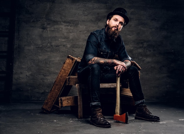 Serious bearded man in hat is sitting with axe in hands.