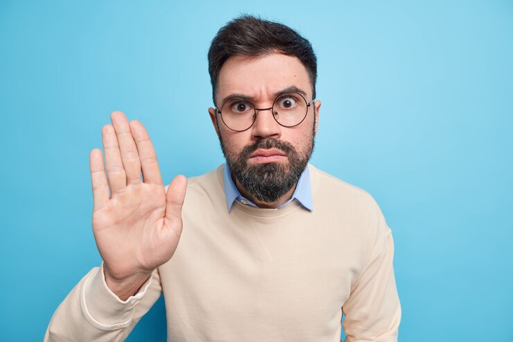 Free Photo serious bearded man keeps palm raised makes restriction or denial gesture wears round transparent glasses casual jumper asks to halt 