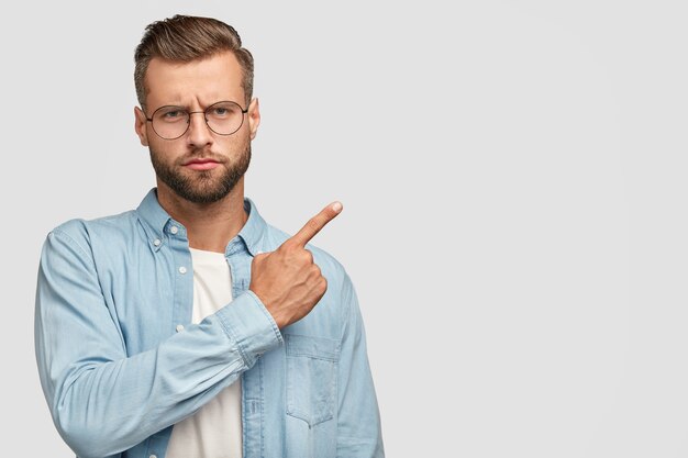 Serious bearded man with strict expression, attracts your attention to something, dressed in blue shirt, shows direction to somewhere