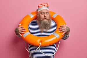Бесплатное фото Серьезный бородатый мужчина в плавательной шапочке и очках смотрит в сторону, носит полосатый матросский жилет, активно проводит летние каникулы, позирует у розовой стены. дежурный спасатель. безопасность пляжного отдыха