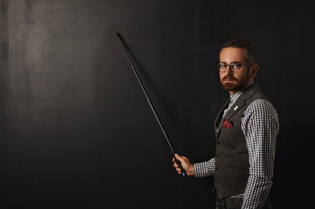 Serious bearded professor in plaid shirt and tweed vest, wearing glasses and looking condemn, shows something on school black board with his pointer