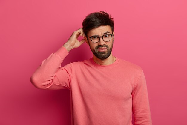 Serious bearded young man scratches head, thinks deeply about how to solve problem