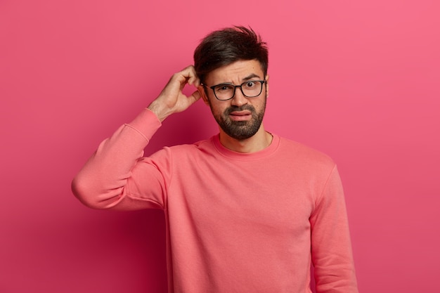 Serious bearded young man scratches head, thinks deeply about how to solve problem