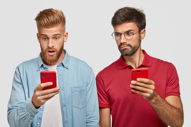 Serious beared male peeks at screen of friend`s smart phone, tries to read message, use modern technologies and high speed internet connection. Stunned red haired hipster uses cellular indoor